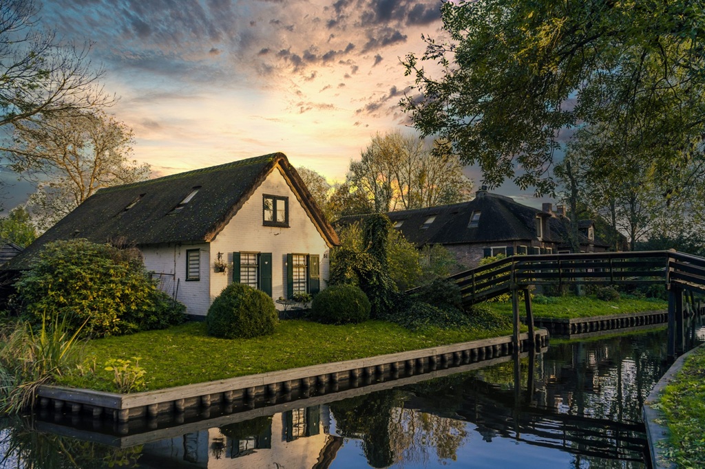 Landelijk huis aan het water met een brug en bomen, bij zonsondergang.
