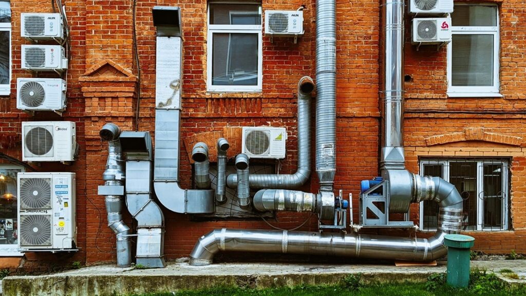 Complexe ventilatie-installatie met luchtkanalen en units aan de buitenzijde van een bakstenen gebouw.
